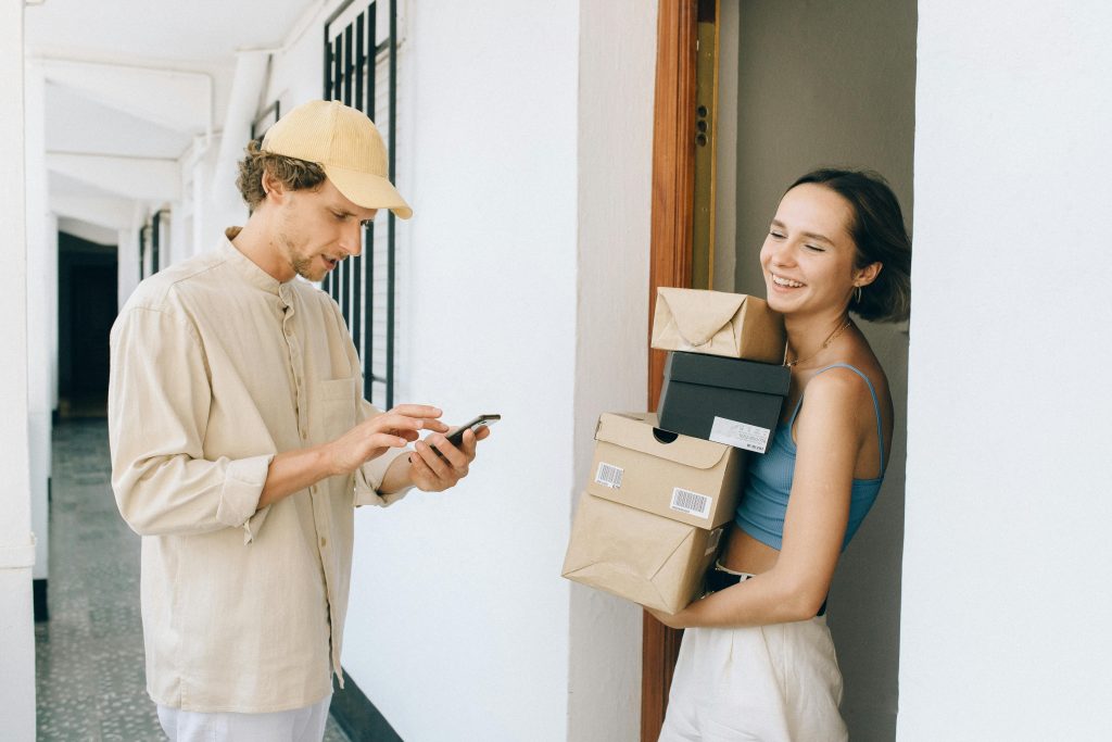 A cheerful woman receives a delivery from a person in a hallway, using a smartphone for online payment.